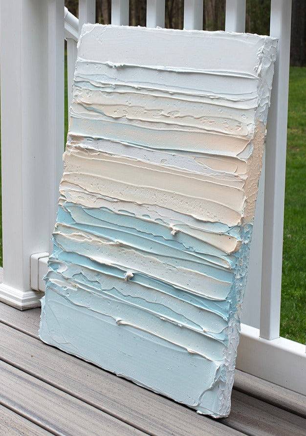 An abstract painting with heavily textured white, sherbet orange and teal blue green paint leaning side view on the artists deck outside.