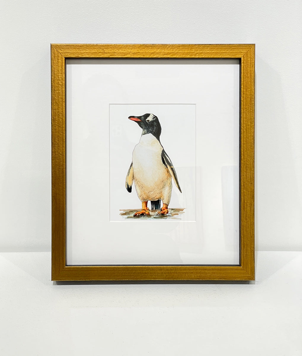 A print of a drawing of a penguin framed in a gold frame with a mat rests on a white surface against a white wall. 