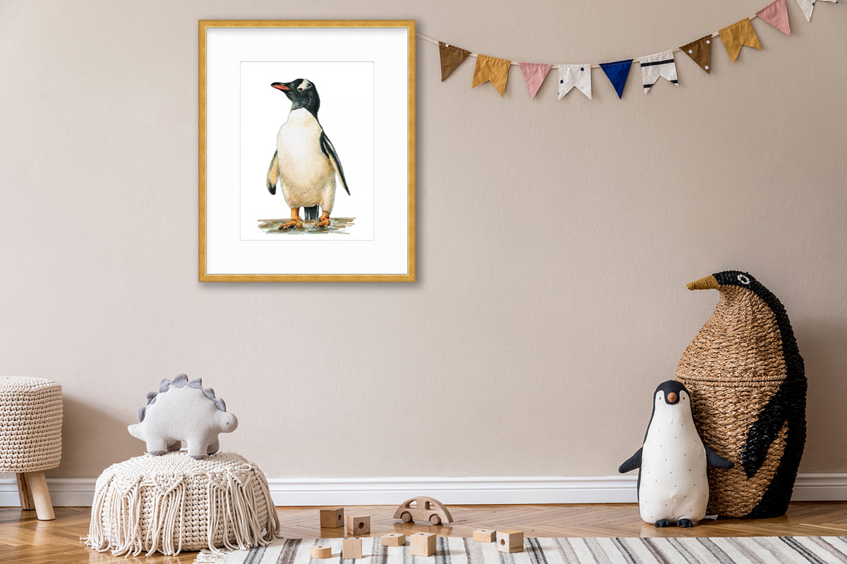 A penguin print in a wood frame with a mat hangs on a beige wall above a white knit ottoman with a stuffed animal dinosaur, wooden blocks and two stand up penguins.