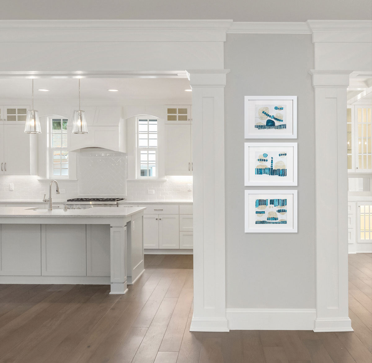 Three abstract blue, beige, and white paintings framed in white frames hang stacked on a grey column wall between the entry to a modern kitchen and the entry to a dining room. 