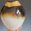 A hand rotates a white, yellow, and umber-toned ceramic vase with a wide gold neck in front of a grey background.