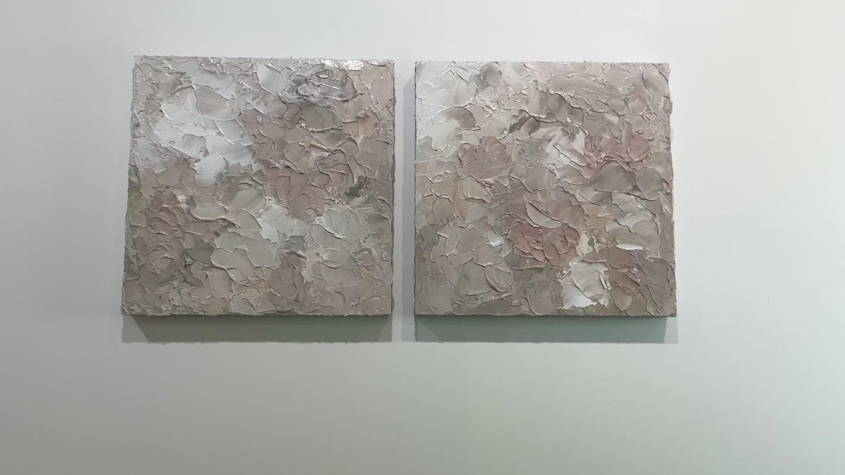 A video of a pair of thickly textured abstract paintings in light pink, grey, coral and white by Teodora Guererra hanging on a white wall.