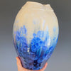 A hand holds a blue and cream-colored glazed ceramic vase with a wide neck in front of a grey backdrop.