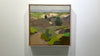 A small framed green and brown landscape painting hangs on a white wall.
