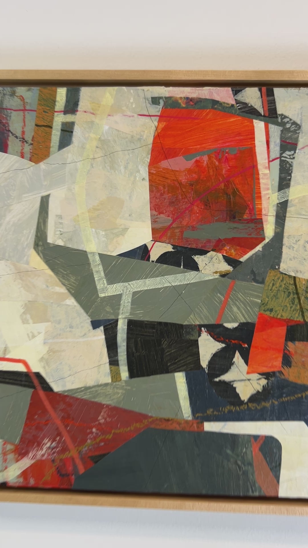 A video of a framed abstract collage artwork featuring geometric shapes in shades of red, beige, and green, with a white background hanging on a white wall.