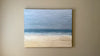 A video of an abstracted coastal seascape painting hangs on a grey wall.