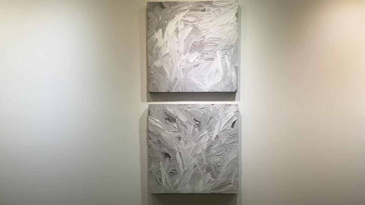A video of a pair of thickly textured paintings in grey, white, blue grey and plum grey by Teodora Guererra hanging on a white wall.