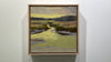 A framed yellow and brown abstracted landscape painting hangs on a white wall.