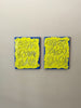 A video of two abstract text based painting featuring a thick, yellow-green impasto texture with blue abstract scribbled lines and a bright blue base hanging on a wall.
