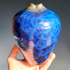 A man's hand holds a vibrant blue glazed ceramic vessel with a silver neck in front of a grey ombre backdrop. 
