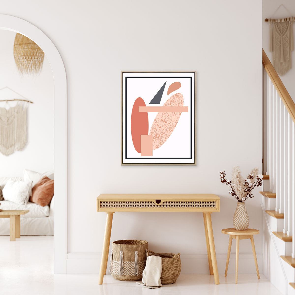 A pink and grey abstract art print framed in a silver frame hangs on a light wall above a console table and between a staircase and an entryway to a living room of a home. 