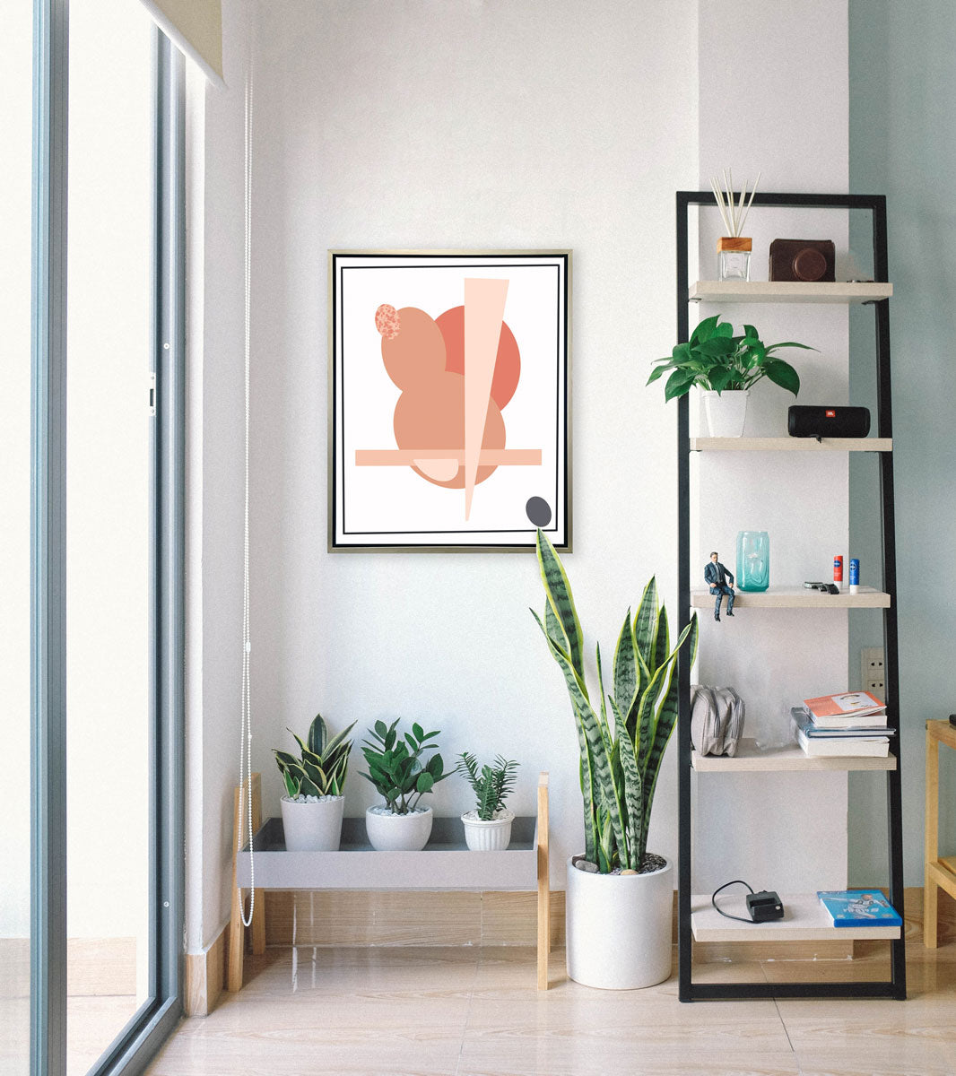 An abstract pink and grey art print framed in a silver frame hangs on a light wall between a floor-to-ceiling window, shelving unit, and potted plants. 