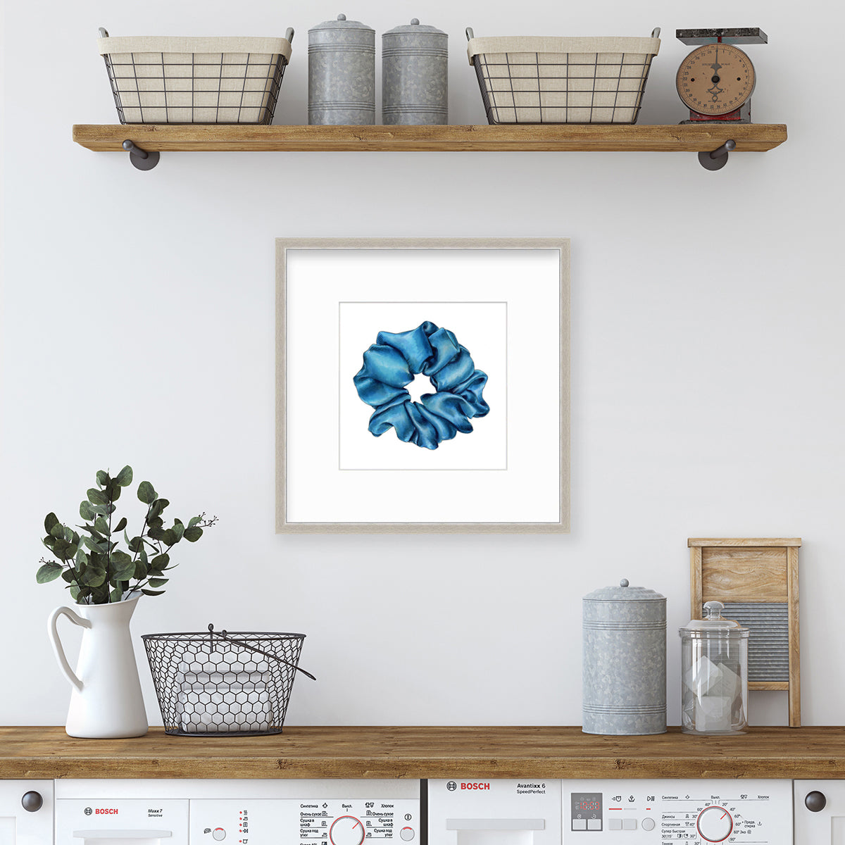 A drawing of a blue hair scrunchie framed in a silver frame hangs on a white wall above a washing unit and below a wall shelf with storage and decor. 
