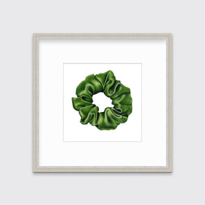 A drawing of a green hair scrunchie framed in a silver frame hangs on a white wall. 