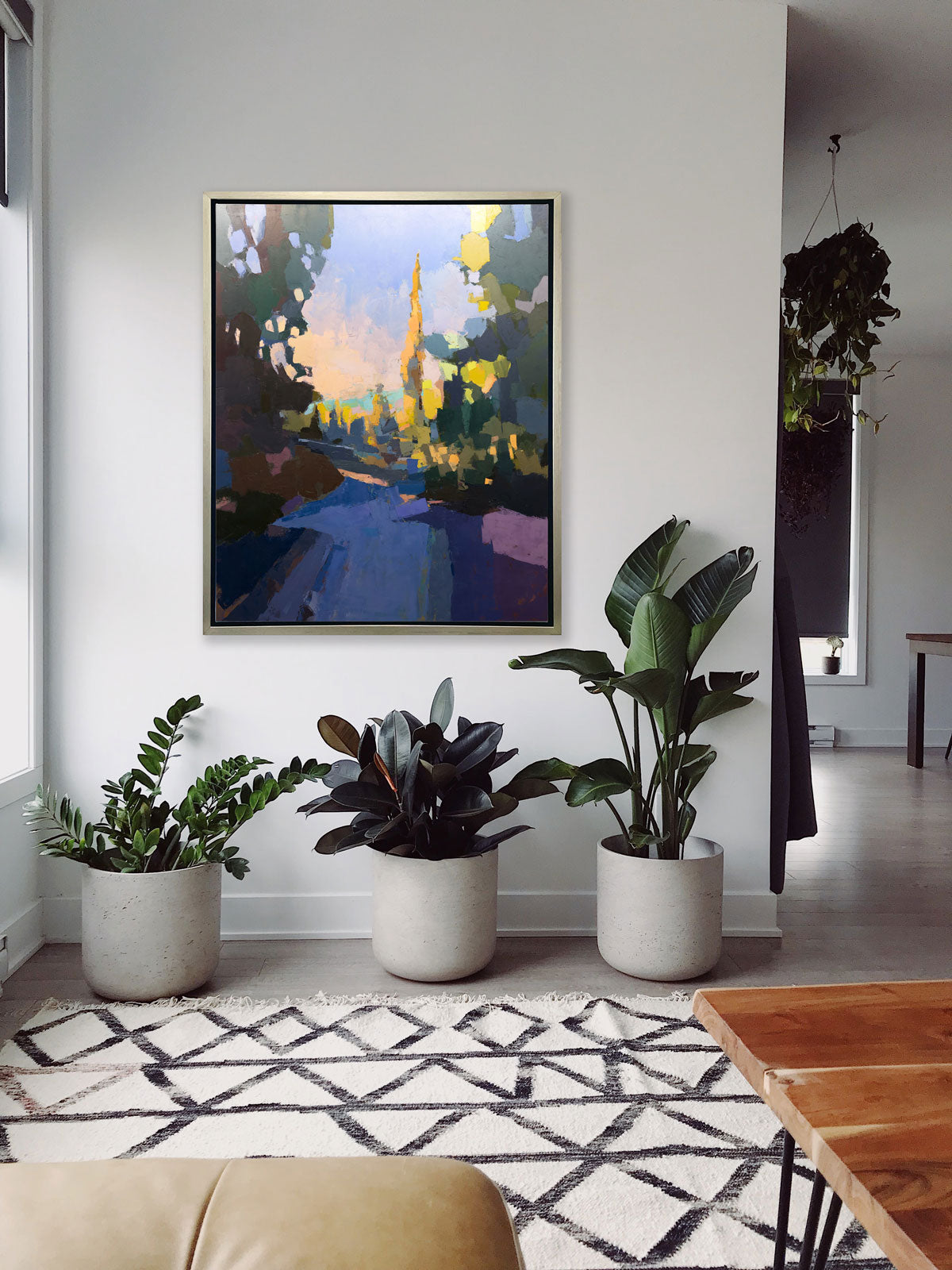 A blue abstract landscape art print framed in a silver frame hangs on a white wall above three large potted floor plants a modern boho rug.