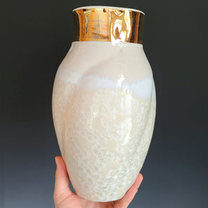 A hand holds a cream-colored glazed ceramic vase with a wide gold neck from it's base in front of a grey back drop. 