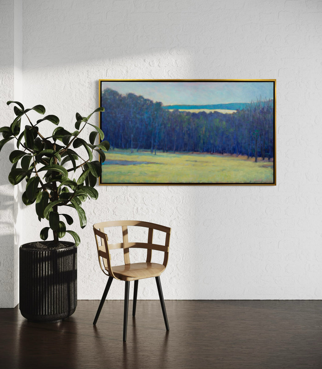 Framed landscape painting on a white wall above a wooden chair and potted plant.