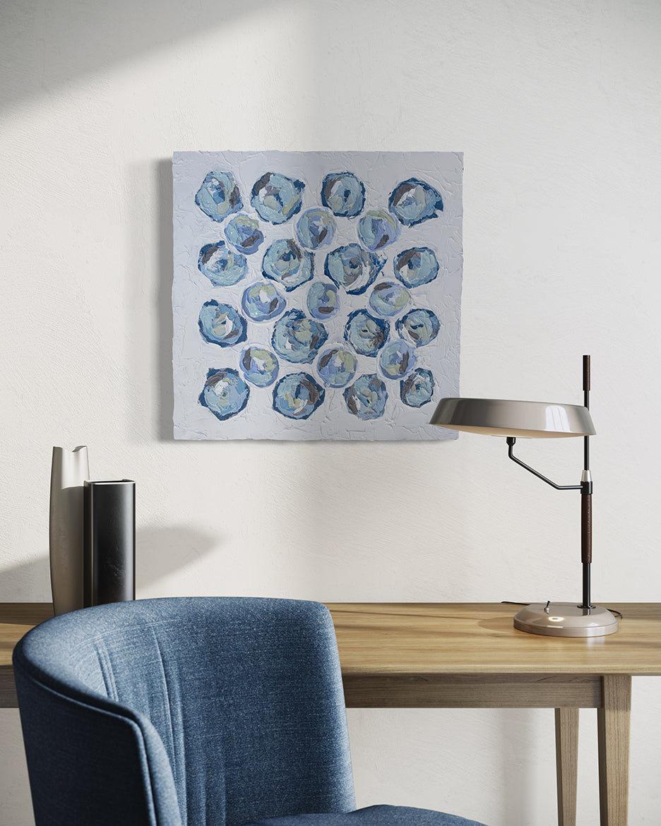 A very textured abstract painting with circle type abstract flowers in blue, teal, green, plum hangs on a white wall above a maple desk with a lamp and vases. A blue chair sits in front of it.