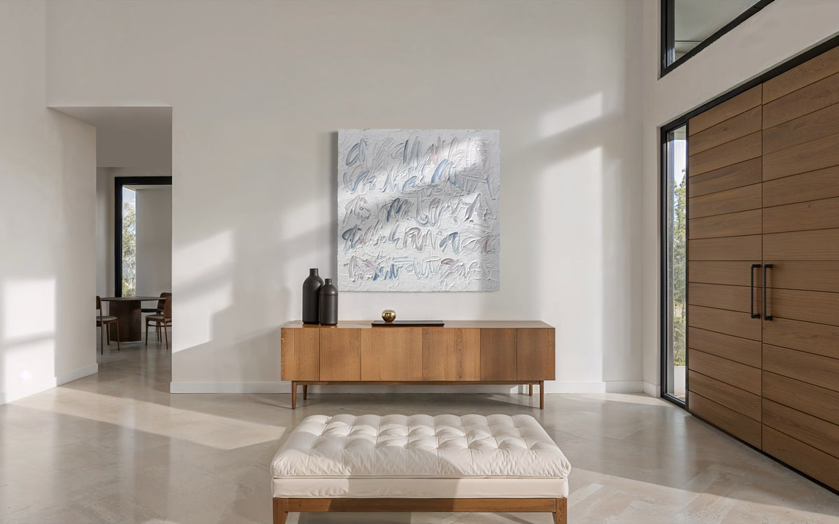 A large square abstract canvas painting with blue and gray text based calligraphic brushstrokes on a white background, displayed on a light wood console table in a modern, sunlit entryway with a tufted bench in the foreground.
