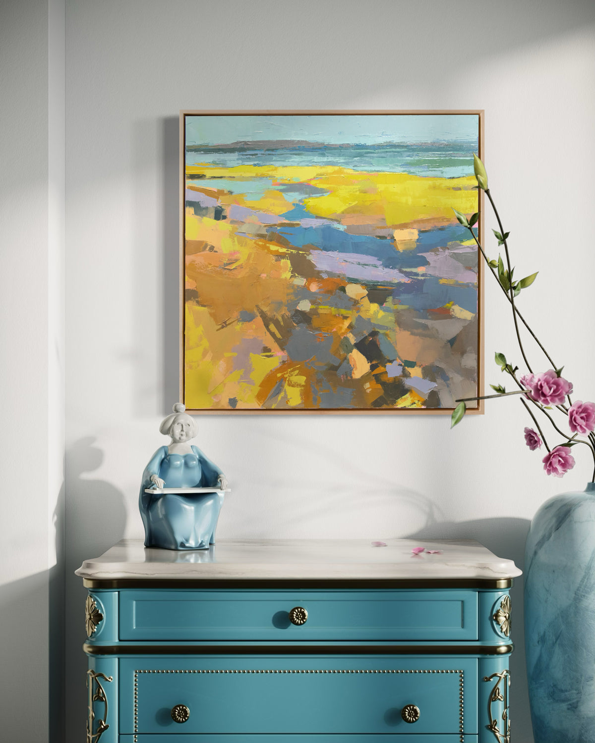 A blue and yellow abstract landscape painting hangs on a white wall over a teal one draw fancy chest with a Buda. 