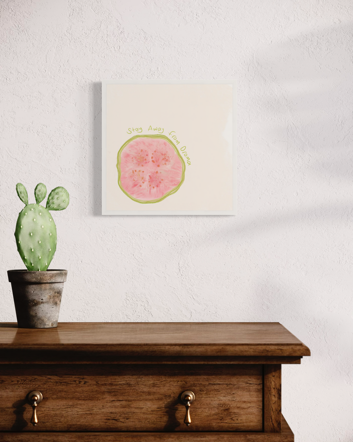 An abstract art print of a fruit with the words "stay away from drama" in an unmatted white frame hangs on a white wall above a stained wood dresser with a small potted cactus.