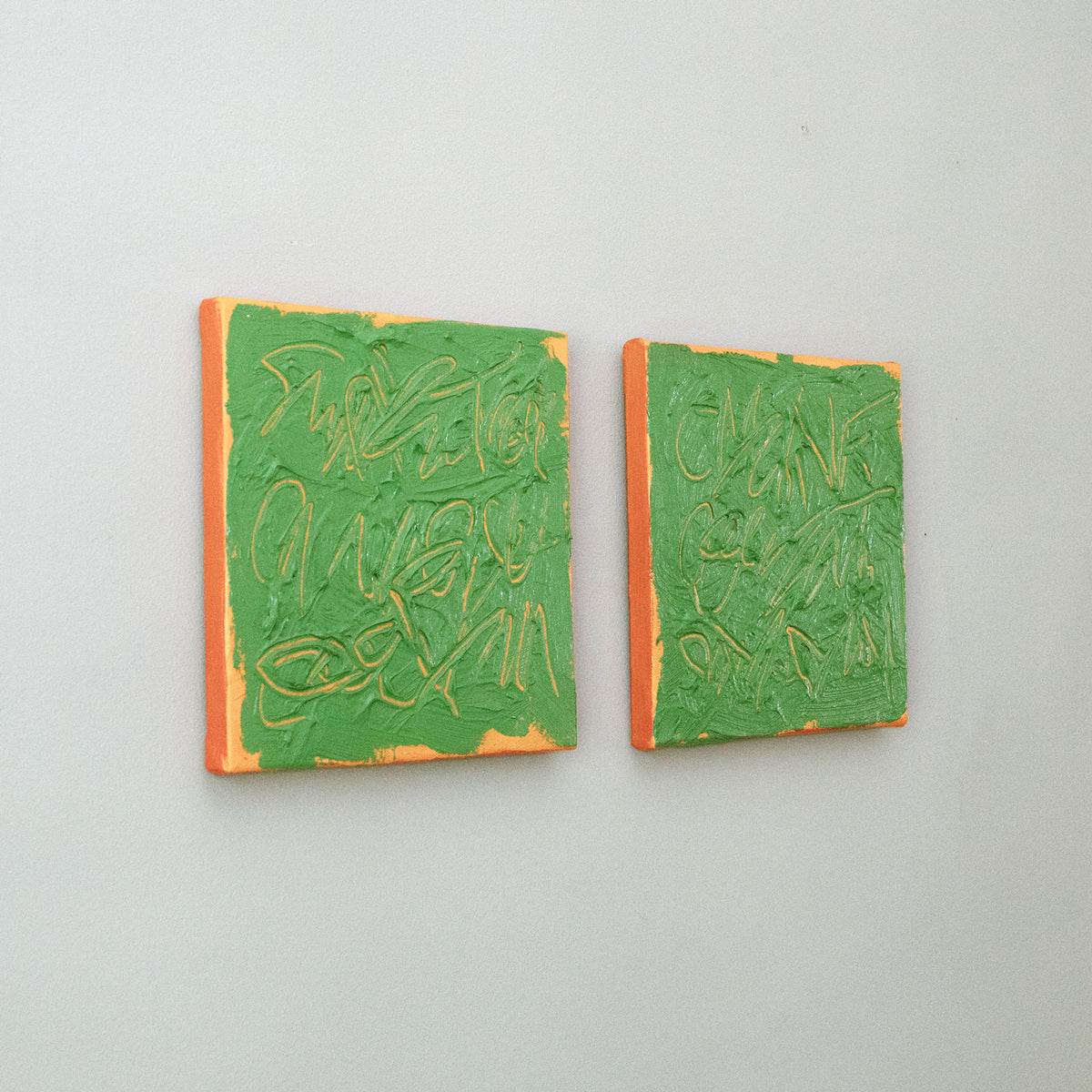 Two square abstract text based paintings with thick green paint and tangerine gestural lines, mounted on a light gray wall.