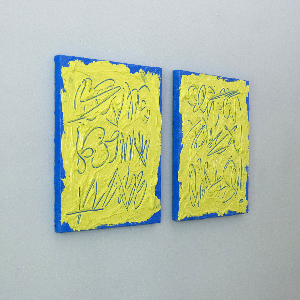 Two abstract text based painting featuring a thick, yellow-green impasto texture with blue abstract scribbled lines and a bright blue background hang on a grey wall, side view.