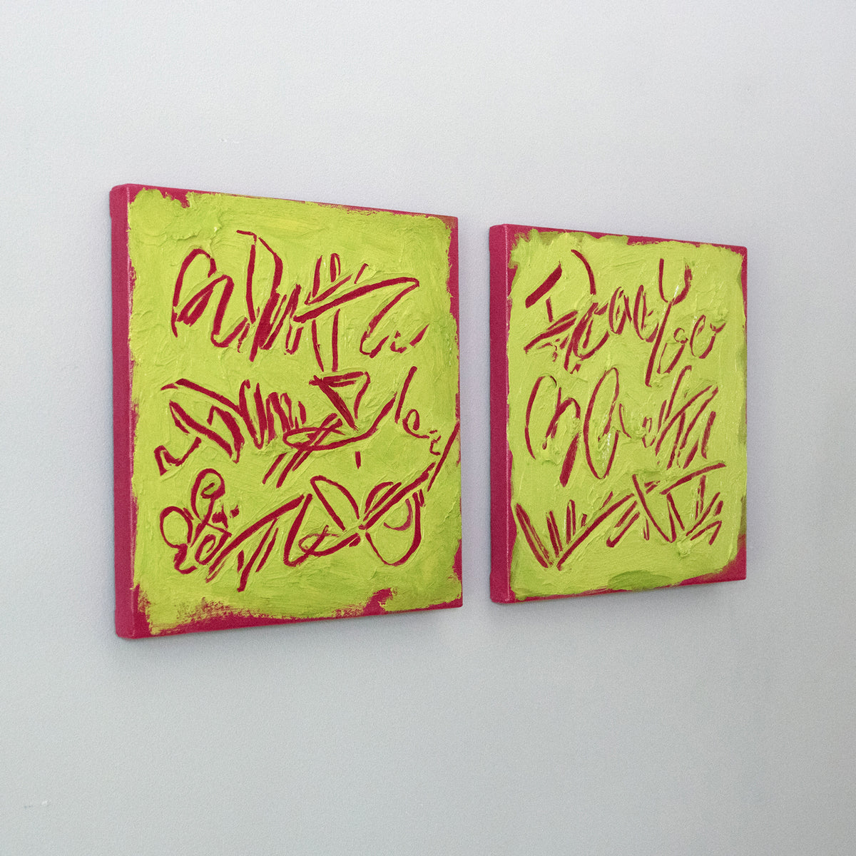 Two square abstract text-based paintings with textured chartreuse over hot raspberry pink  graffiti-style lettering, displayed side-by-side on a white wall, side view.