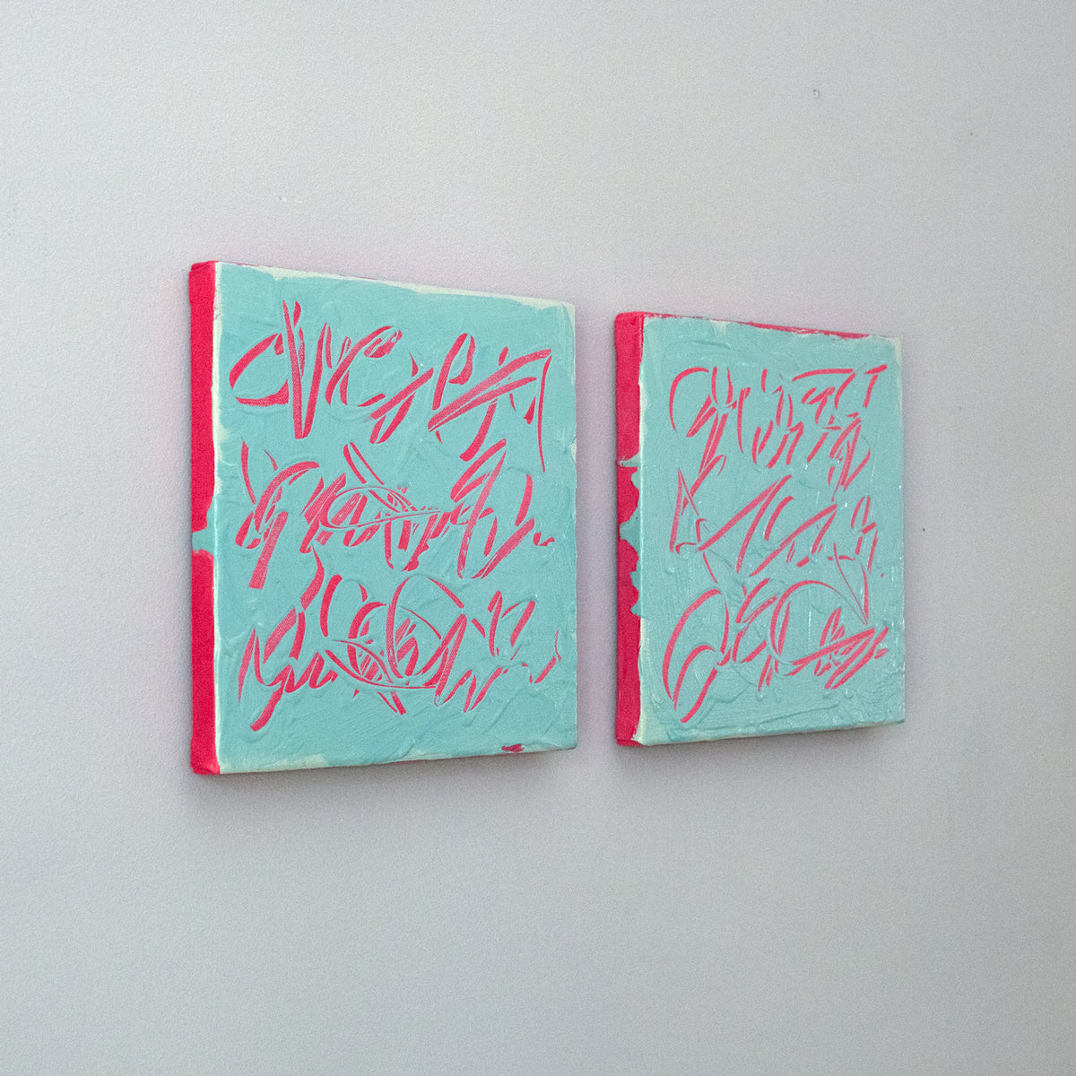 Two square abstract text based paintings with turquoise backgrounds and pink scribbled lines, displayed side by side on a light gray wall, side view.