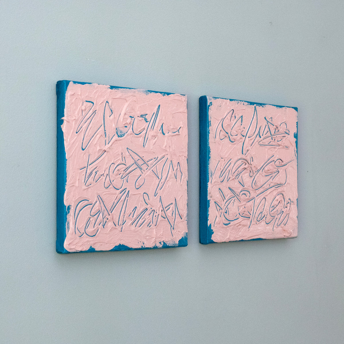 Two square text based paintings with textured pink surfaces and blue edges, featuring abstract teal line drawings, displayed side by side on a light blue-gray wall.