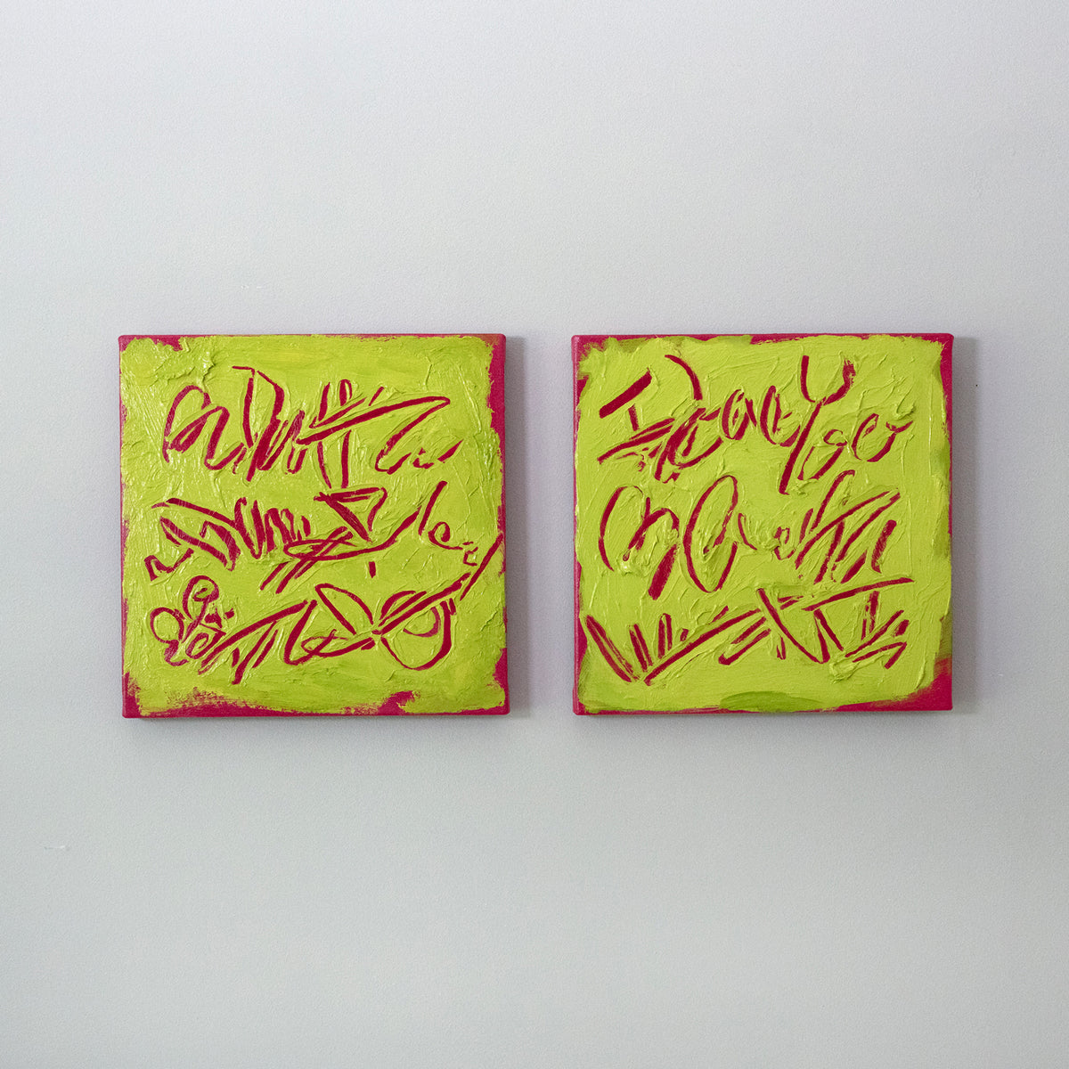 Two square abstract text-based paintings with textured chartreuse over hot raspberry pink  graffiti-style lettering, displayed side-by-side on a white wall.