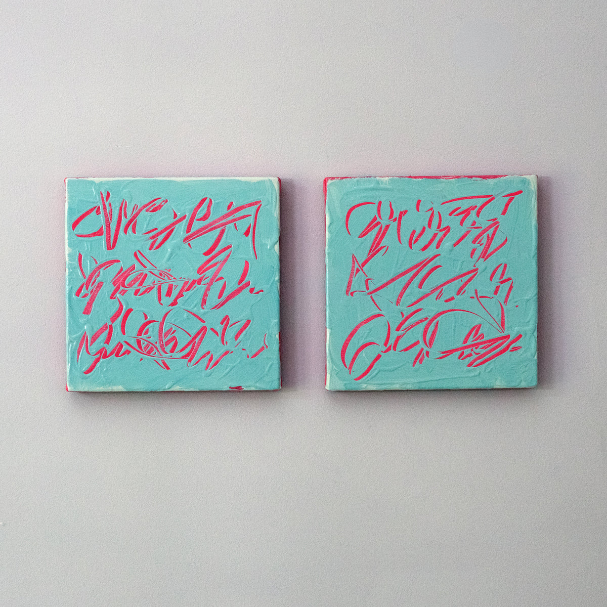 Two square abstract text based paintings with turquoise backgrounds and pink scribbled lines, displayed side by side on a light gray wall.