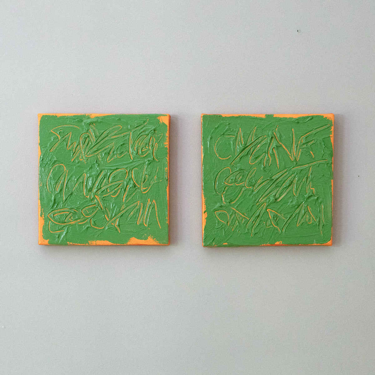 Two square abstract paintings side by side on a light gray wall. Each painting features textured green paint with tangerine texted abstract brushstrokes and a thin orange border.