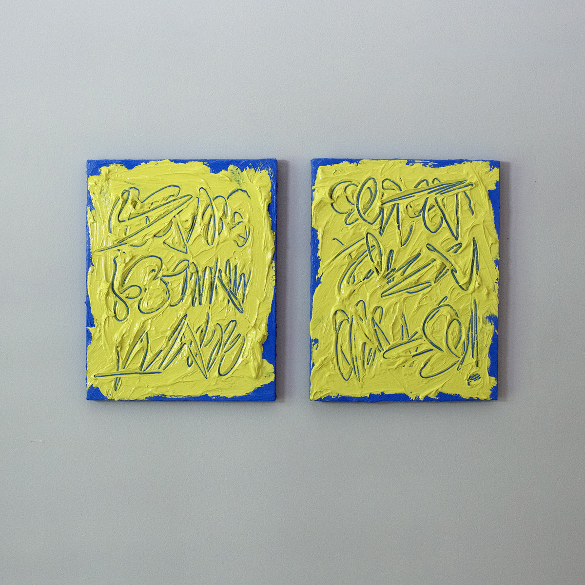 Two abstract text based painting featuring a thick, yellow-green impasto texture with blue abstract scribbled lines and a bright blue background hang on a grey wall.