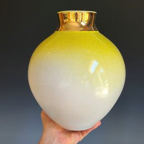 A hand holds a glazed ceramic vessel which is white at the bottom and fades to bright yellow at the top, with a wide gold neck in front of a grey background. 