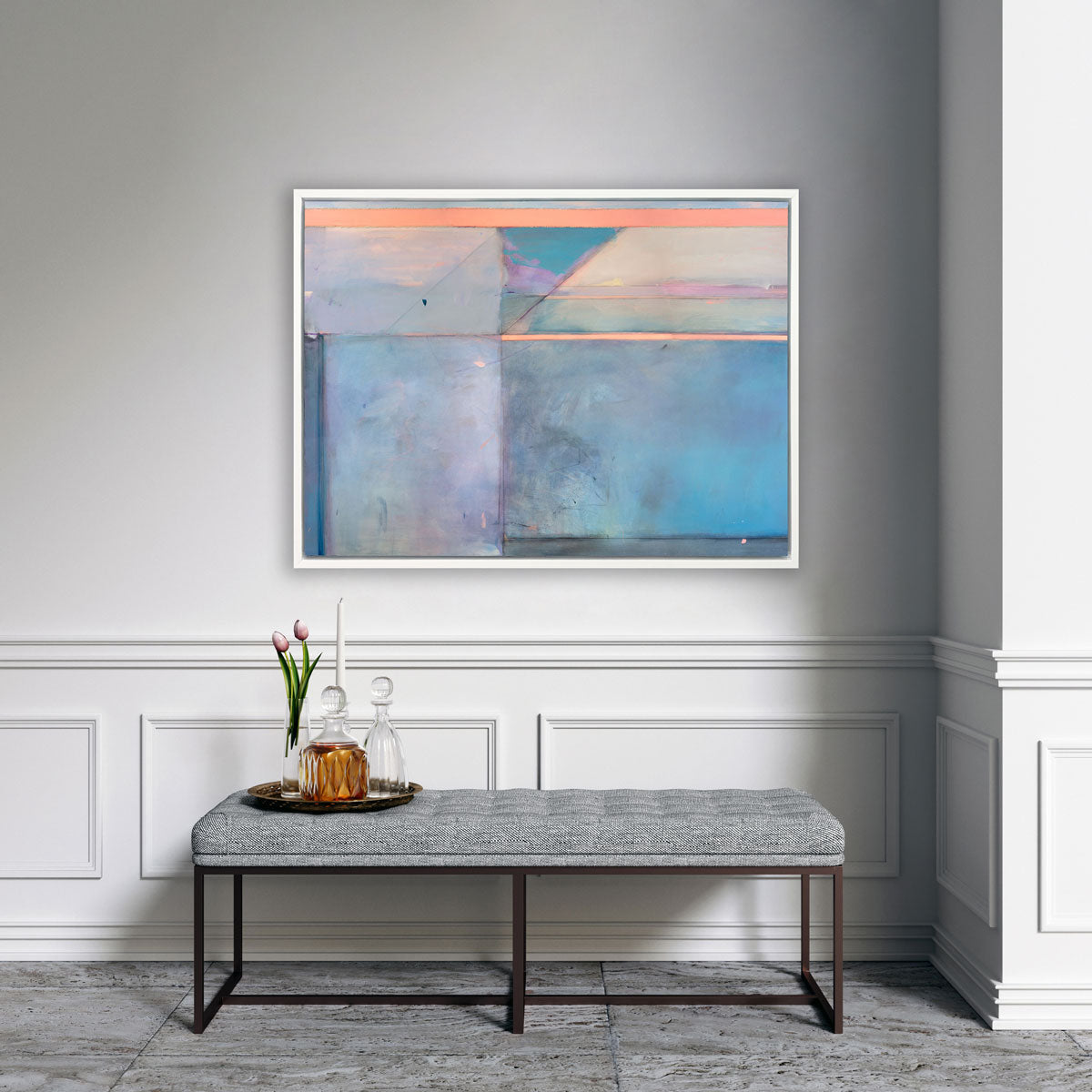 A pink and blue abstract print by Kelly Rossetti framed in a white floater frame hangs on a white wall above a grey bench with a tray with a two decanters, a candle, and tulips in a glass vase. 