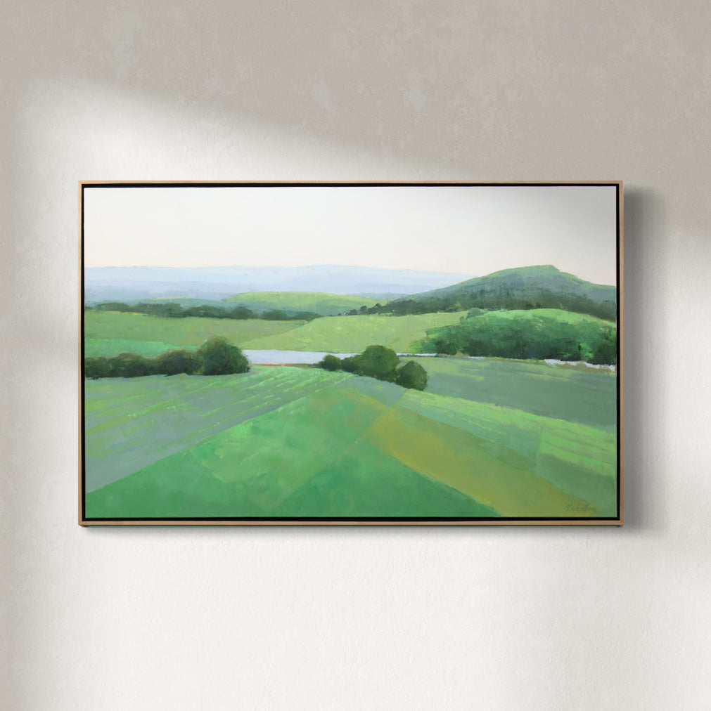 A green landscape painting framed in a light wood floater frame hangs on a white wall.