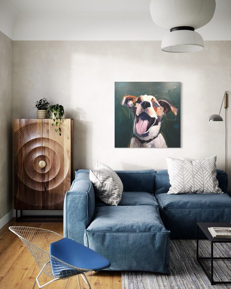 A square painting depicting a close-up portrait of a white and brown dog with its tongue out, displayed on a beige wall above a blue fabric sectional sofa in a modern living room setting.