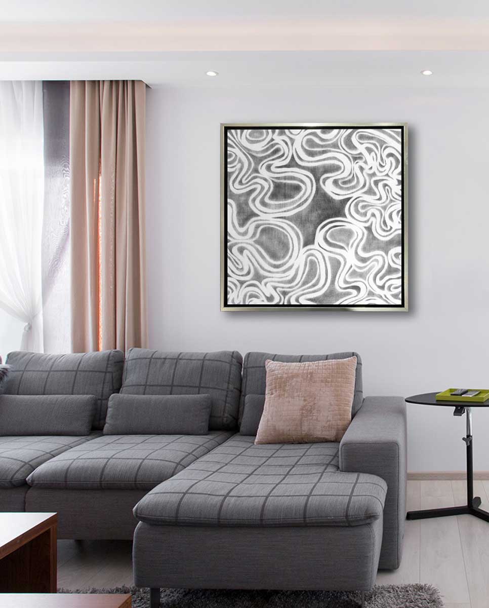 A grey and white abstract print in a silver floater frame hangs on a white wall above a grey sofa with a light pink pillow and a small black pedestal side table.