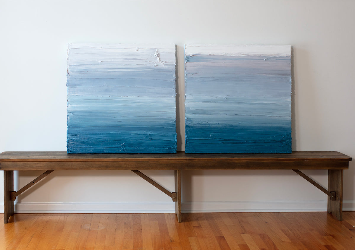 Two white, light grey and tonal blue abstract paintings by Teodora Guererra rest on a wood bench against a white wall.