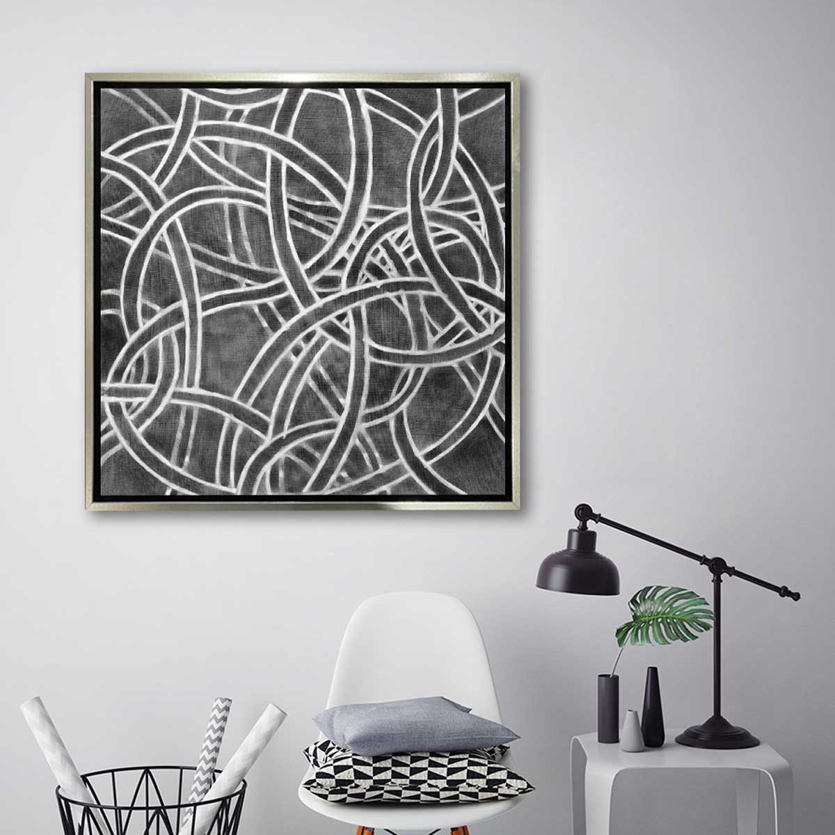 A dark grey and white abstract print in a silver floater frame hangs on a white wall above a white chair with pillows, a small white side table with a black desk lamp and a black wire basket holding printed wrapping paper.