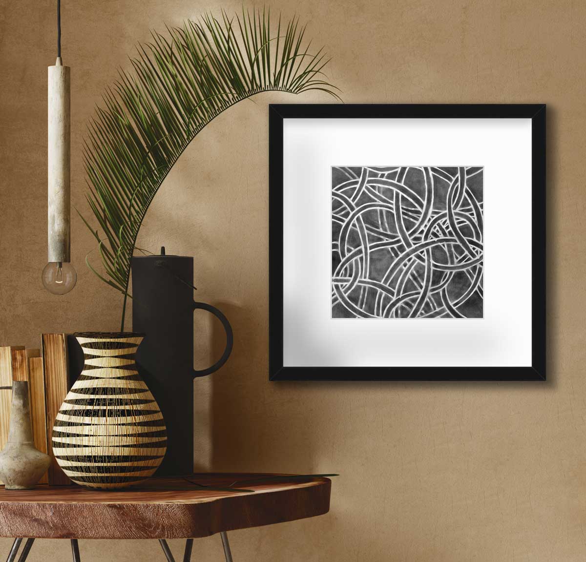 A dark grey and white abstract print in a black frame with a mat hangs on a beige wall next to a live edge side table with a black cylindrical pitcher, books and a beige and black vase with a large leaf stem.