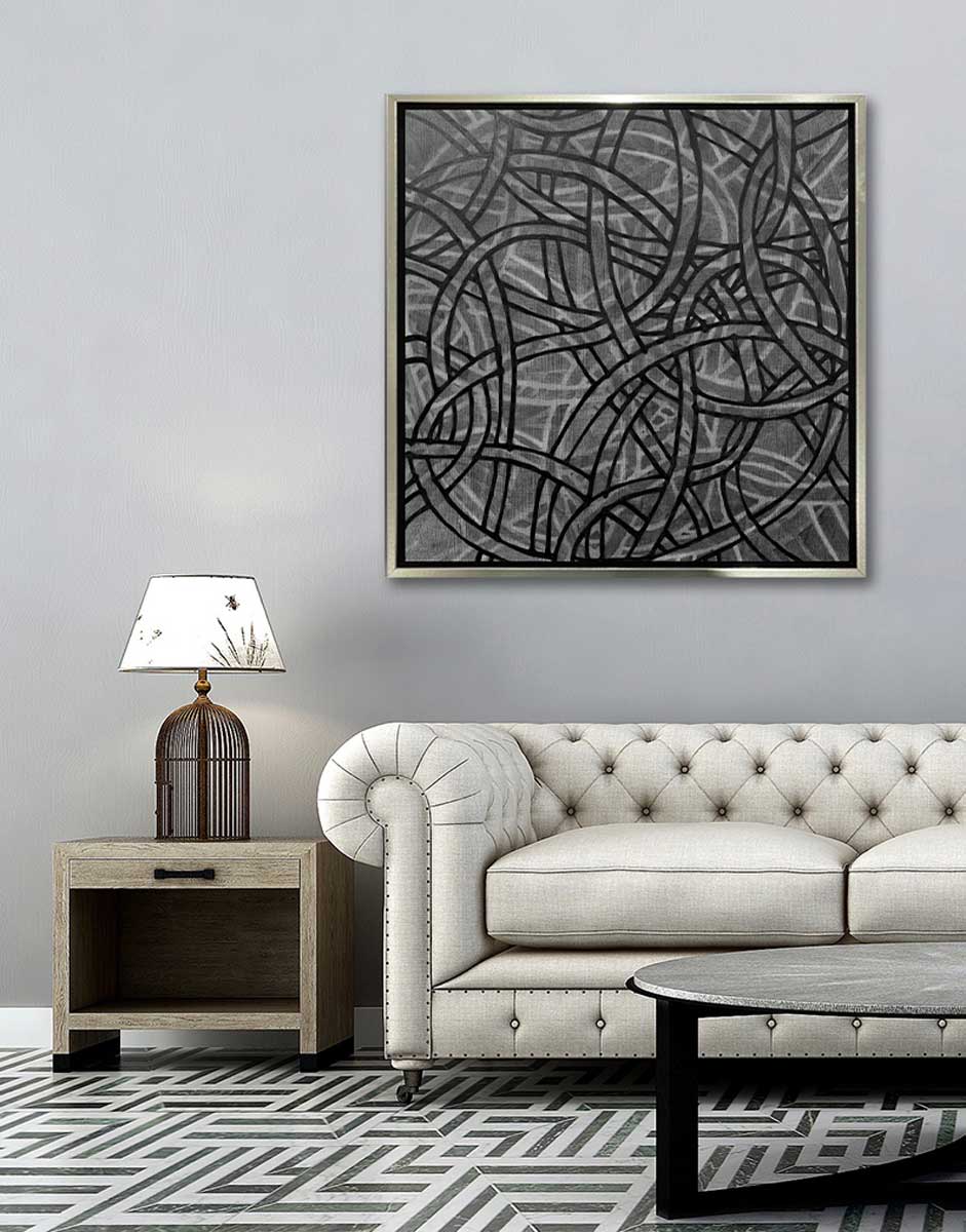 A grey, black and white abstract print in a silver floater frame hangs on a white wall above a cream tufted sofa, a round black and white coffee table and a natural wood side table with a birdcage table lamp.