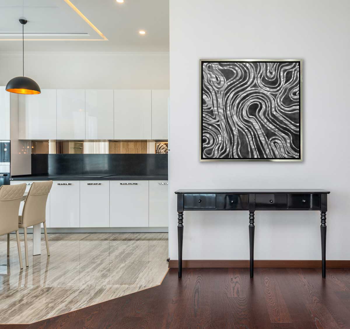 A dark grey and white abstract print in a silver floater frame hangs on a white wall above a black console table and next to a kitchen with black countertops, white cabinets and beige chairs.