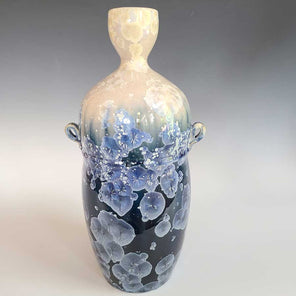 A white, cream, dark blue and light blue glazed narrow vase with two small handles sits on a white surface.