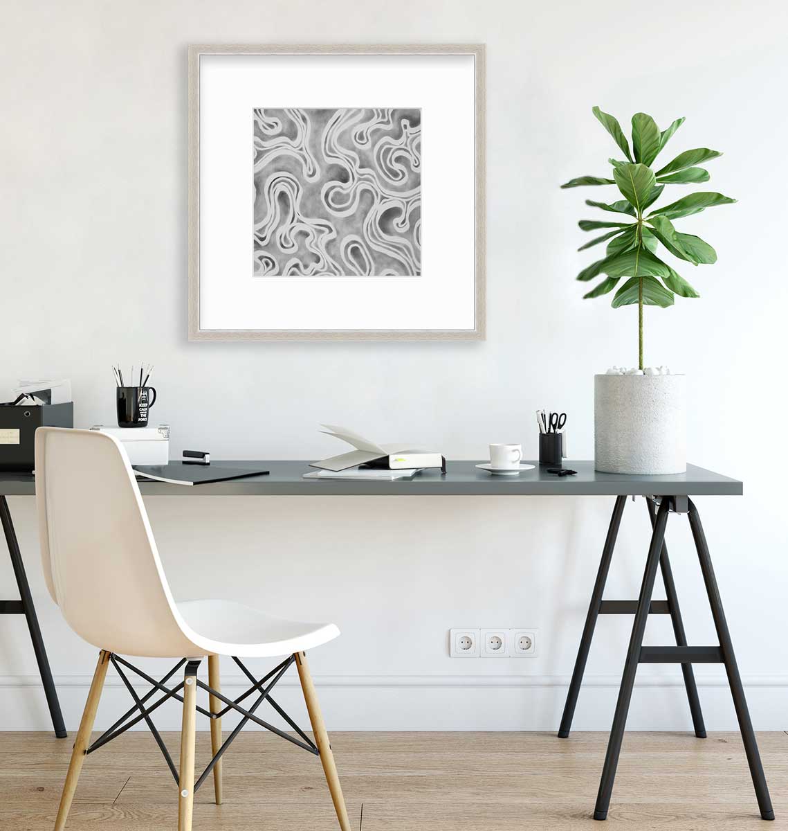A grey and white abstract print in a silver frame with a mat hangs on a white wall above a grey desk with an open notebook, coffee mugs with pencils and a potted plant.