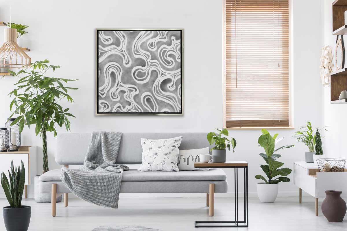 A grey and white abstract print in a silver floater frame hangs on a white wall above a grey modern sofa with printed pillows, several potted floor plants and a wood and iron side table.