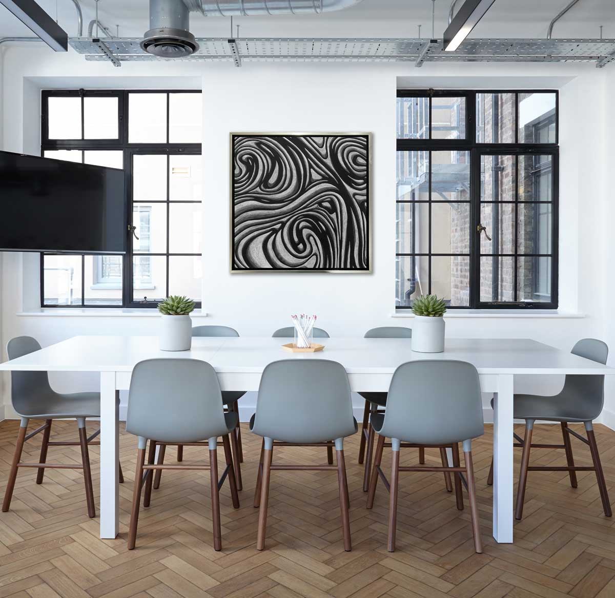 A black and grey abstract print in a silver floater frame hangs on a white wall in between two large windows and above a long white table surrounded by eight grey chairs.