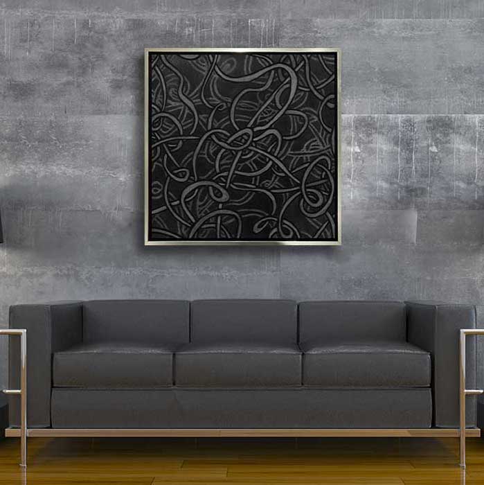 A black and dark grey abstract print in a silver floater frame hangs on a grey industrial wall above a modern grey and chrome sofa.