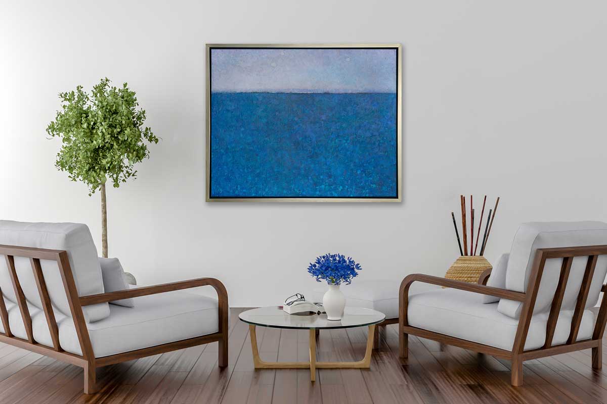 A deep blue abstract landscape print in a silver floater frame hangs on a white wall above two oversized wood chairs with white cushions and pillows, a small round coffee table with a white vase with periwinkle flowers and a white matching ottoman.